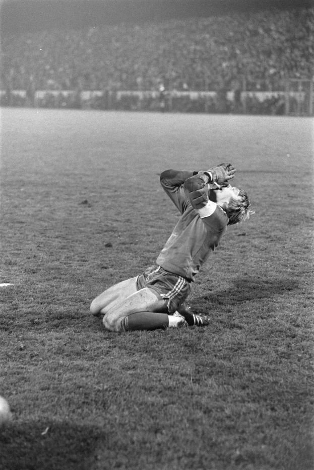 Ein Schwarz-Weiß-Foto eines Fußballspielers, der auf dem Boden kniet und seinen Kopf in den Händen hält, umgeben von einer Menschenmenge im Hintergrund.