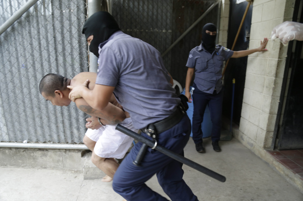 Ein Mann in Gewahrsam, der von zwei Polizeibeamten vor einem Gebäude eskortiert wird, wobei einer der Beamten seinen Arm hält und der andere einen Schlagstock hält, neben einer Wand, einer Tür, einem Rohr und einem Maschendrahtzaun.