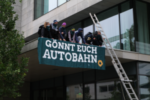Eine Gruppe von Menschen auf dem Dach eines Gebäudes, die eine Fahne halten, mit einem Baum links und Glasfenstern im Hintergrund, wahrscheinlich protestierend im Namen der deutschen Regierung.