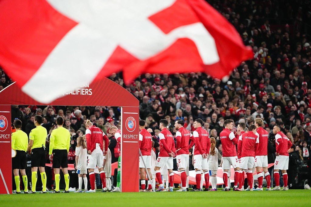 Gruppe von Menschen auf einem Fußballfeld mit einer roten und weißen Flagge im Vordergrund, einem Bogen mit der Aufschrift "Bayern München vs. Bayern München Wetten und Vorschau" im Hintergrund und einer großen Menge im Stadion.