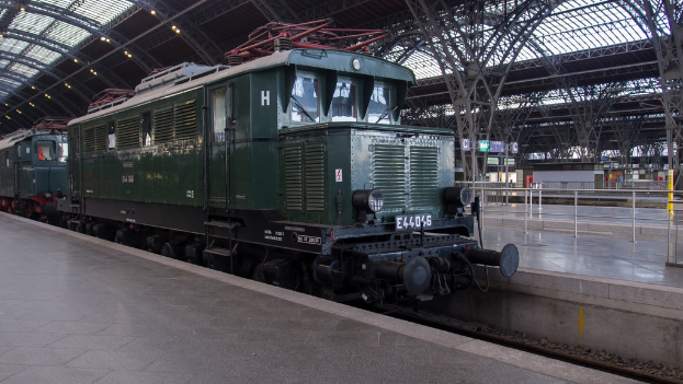 Ein Zug auf Schienen mit Geländern und Brettern im Hintergrund, Lampen auf dem Dach und Himmel darüber, sowie ein Boden darunter.