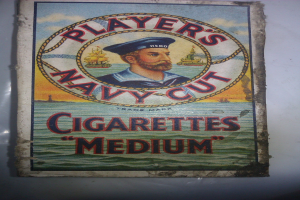 Plakat, das "Player's Navy Cut Cigarettes Medium" bewirbt, zeigt eine Person in einer marineblauen Uniform mit weißem Hemd und roter Krawatte, die Arme verschränkt und mit ernstem Gesichtsausdruck, sowie fetten schwarzen Text in einem weißen Rahmen.