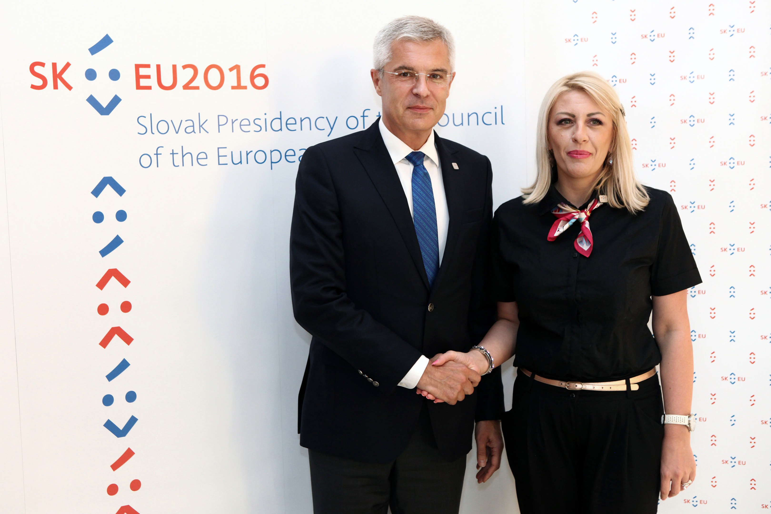 Zwei Personen, ein Mann in einem schwarzen Blazer und weißem Hemd mit blauer Krawatte und eine Frau mit Brille, stehen strahlend nebeneinander vor einer Wand mit einer Whiteboard, auf dem "sk eu 2016" steht, identifiziert als Präsident des Europäischen Rates.