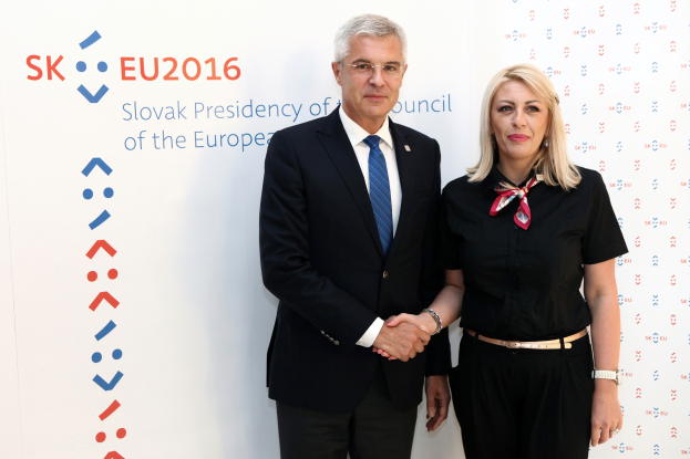 Zwei Personen, ein Mann in einem schwarzen Blazer und weißem Hemd mit blauer Krawatte und eine Frau mit Brille, stehen strahlend nebeneinander vor einer Wand mit einer Whiteboard, auf dem "sk eu 2016" steht, identifiziert als Präsident des Europäischen Rates.