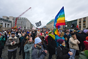 Eine große Gruppe von Menschen steht vor einem Gebäude und hält Fahnen und Schilder mit der Aufschrift "LGBTQ+ rights march in Berlin", mit Gebäuden, einem Kran und einem bewölkten Himmel im Hintergrund.