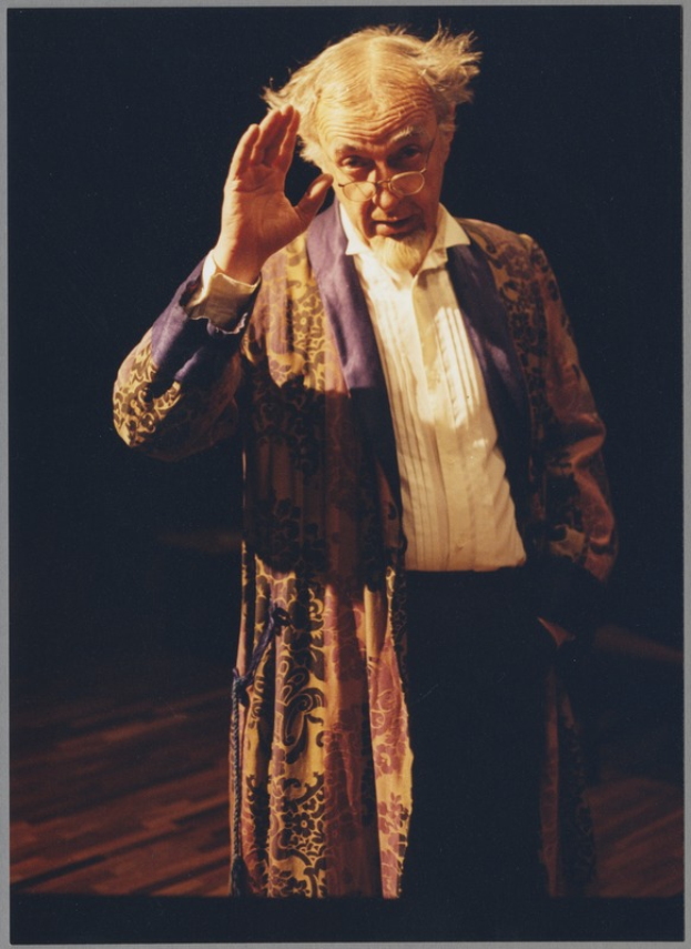 Ein Mann in einem Gewand und Brille steht auf einer Bühne mit dunklem Hintergrund und schaut ernsthaft ins Publikum.