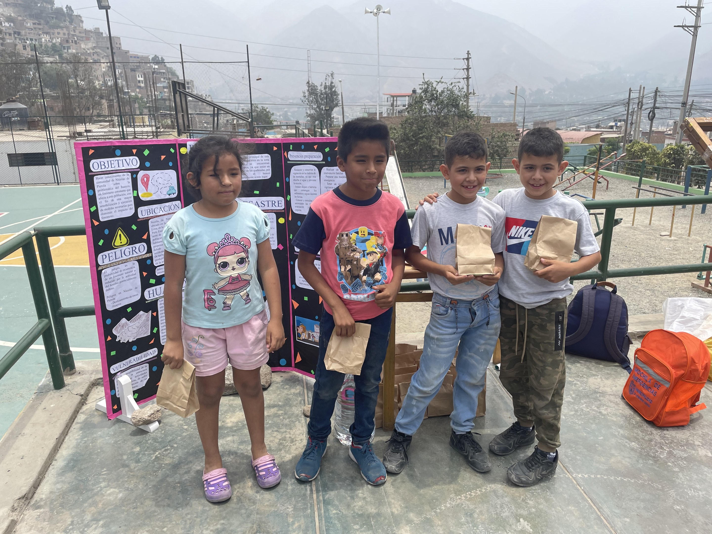 Gruppe von Kindern auf einem Basketballfeld mit Papiertüten in den Händen, Tüten und Gegenstände rechts und eine Tafel mit Text links, vor einer Kulisse aus Zaun, Pfosten, Straßenlaternen, Bäumen, Gebäuden, Hügeln und Himmel.