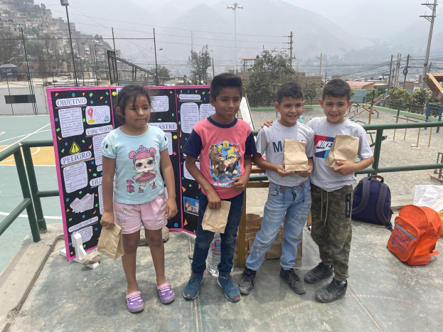 Gruppe von Kindern auf einem Basketballfeld mit Papiertüten in den Händen, Tüten und Gegenstände rechts und eine Tafel mit Text links, vor einer Kulisse aus Zaun, Pfosten, Straßenlaternen, Bäumen, Gebäuden, Hügeln und Himmel.