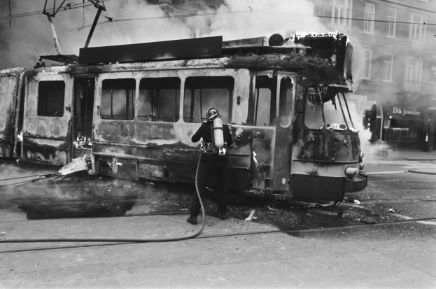 Schwarz-weißes Bild eines Mannes in einem Helm, der eine Pfeife h├Ąlt und neben einer ausgebrannten Stra├čenbahn mit aufsteigendem Rauch steht, auf einer Stra├če mit einem Geb├Ąude und Kabeln im Hintergrund.