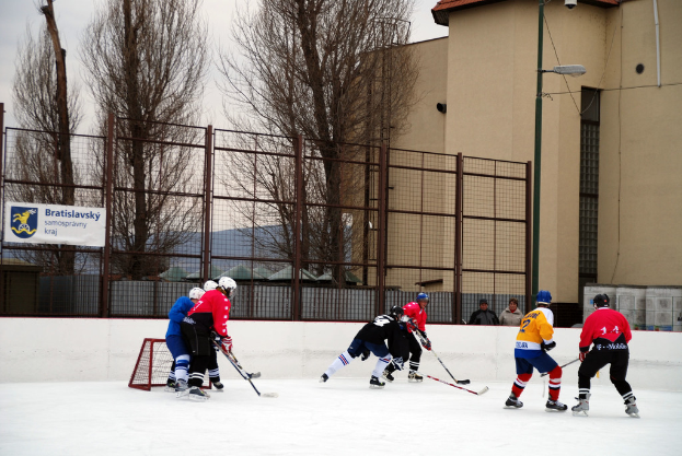 Personen spielen Eishockey auf einem Eis mit Gebäuden, Bäumen, einer Straßenlaterne, einem Namensschild und Zäunen im Hintergrund unter einem klaren Himmel.