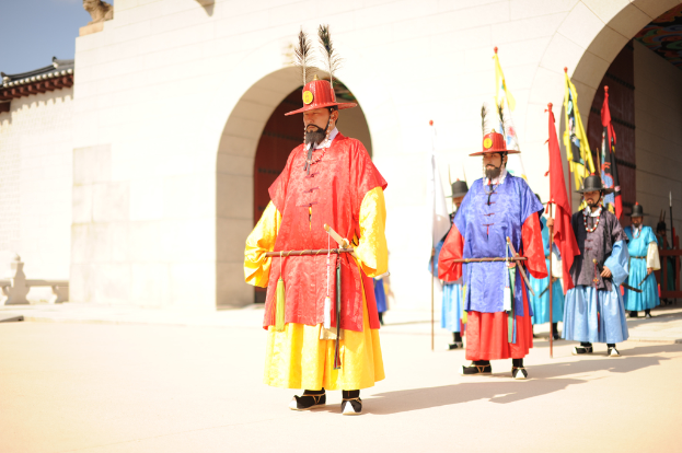 Eine Gruppe von Menschen, die draußen stehen, einige mit Hüten, Fahnen und Speeren, mit einem Gebäude im Hintergrund.