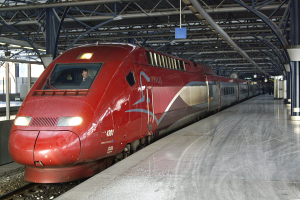 Ein roter Hochgeschwindigkeitszug steht auf einem Bahnsteig mit einigen Menschen und Säulen und Stangen im Hintergrund.