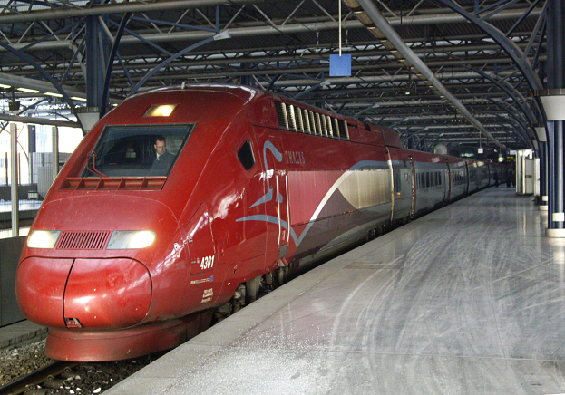 Ein roter Hochgeschwindigkeitszug steht auf einem Bahnsteig mit einigen Menschen und Säulen und Stangen im Hintergrund.