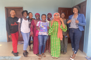 Eine Gruppe von Frauen, die vor einem Schulgebäude in Nigeria lächeln und Bücher halten, mit einem Tuch auf dem Boden und sichtbaren Türen im Hintergrund.