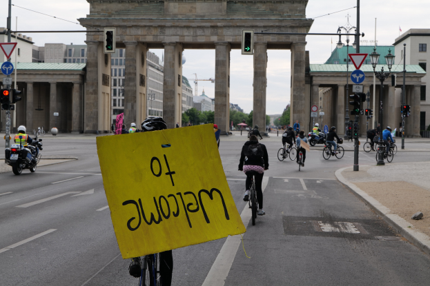 Eine Gruppe von Radfahrern fährt am Brandenburger Tor in Berlin, Deutschland vorbei, einer hält ein gelbes Schild; Laternenpfähle und Verkehrszeichen säumen die Straße unter einem klaren blauen Himmel.