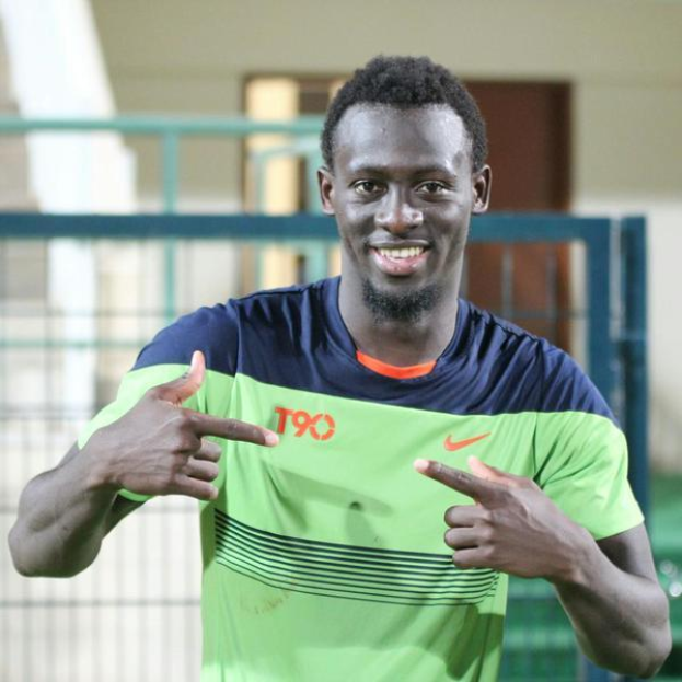 Ein Mann in einem grün-blauen Hemd, wahrscheinlich Emmanuel Adebayor, zeigt mit dem Finger auf die Kamera und lächelt, mit einem unscharfen Geländer, einer Wand und einer Tür im Hintergrund.