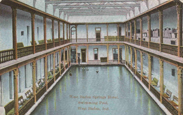 Schwarz-weiß-Postkarte eines Innen-Schwimmbads im West Baden Springs Hotel mit schwimmenden Menschen, umgeben von architektonischen Details und Text am unteren Rand.