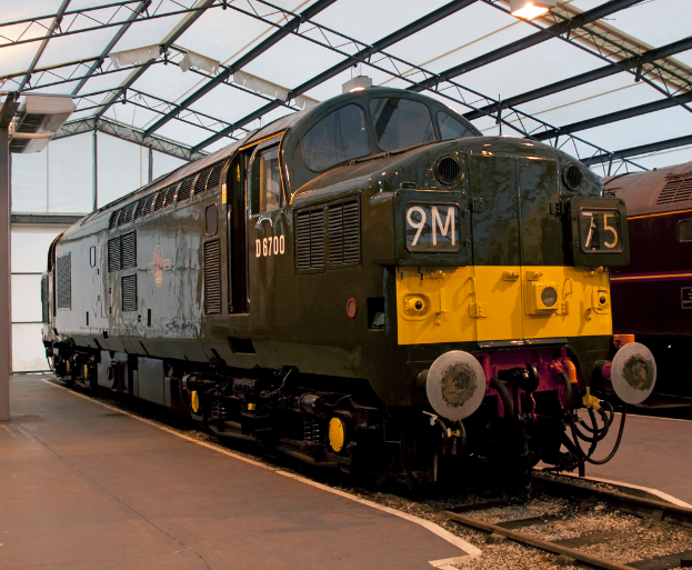 Ein Zug fährt auf den Schienen neben einer Plattform mit einem an der Decke befestigten Pfosten, der Metallstangen mit Lampen trägt.