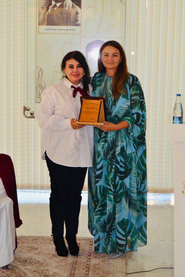 Zwei Frauen auf einer Preisverleihung, eine hält einen Preis, mit einem Rednerpult, einer Wasserflasche, Vorhängen und einem Foto im Hintergrund.