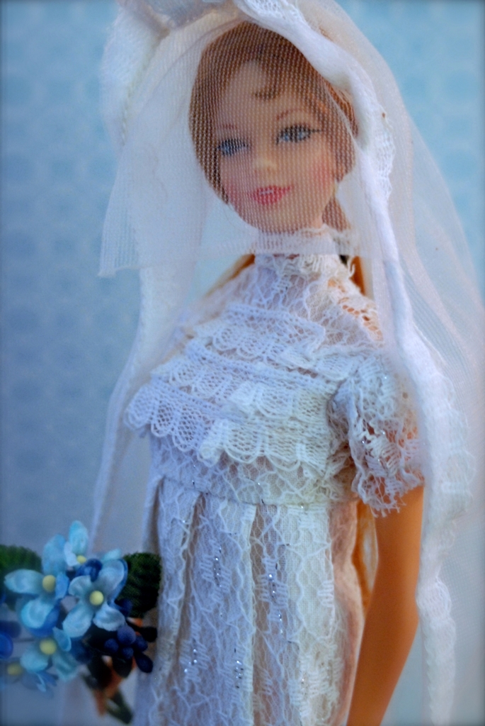 Eine Puppe in einem Kleid, Schleier und mit einem Blumenstrauß, vor einem unscharfen Objekt.