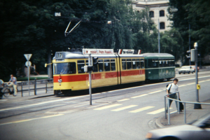 Ein altes Foto einer gelben und roten Straßenbahn auf einer Stadtstra{\ss}e mit Fahrzeugen, Fussgängern, Fahrradfahrern, Bäumen und einem Gebäude im Hintergrund.