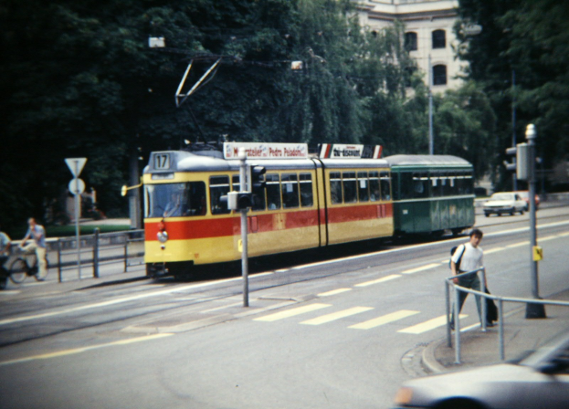 Ein altes Foto einer gelben und roten Straßenbahn auf einer Stadtstra{\ss}e mit Fahrzeugen, Fussgängern, Fahrradfahrern, Bäumen und einem Gebäude im Hintergrund.