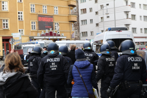 Polizeibeamte in Uniform vor einer Menge bei einer Demonstration in Berlin, mit Fahrzeugen, Gebäuden und einer Person mit einer Kamera.