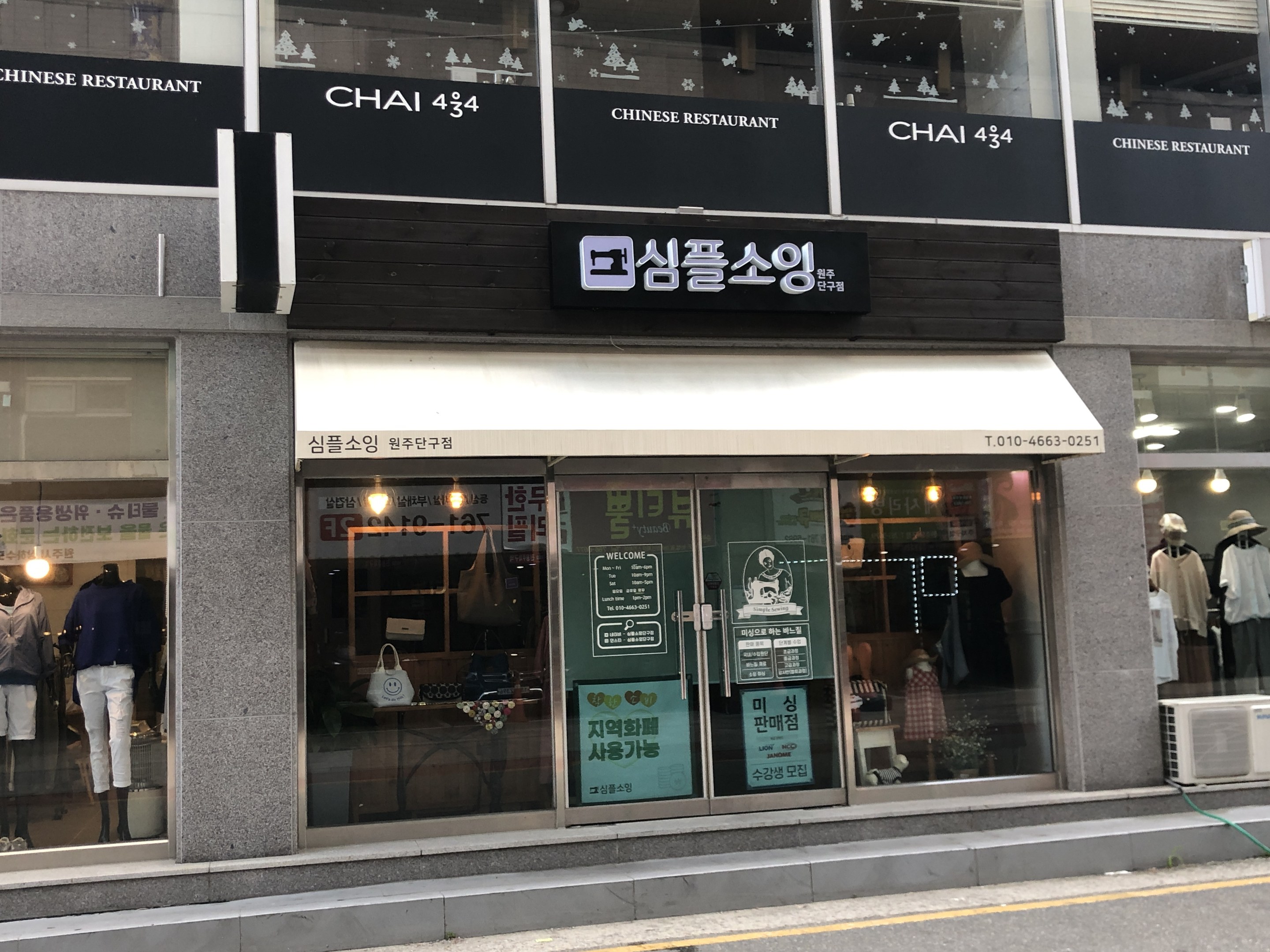 Außensicht des Chai 434 Chinesischen Restaurants in Seoul, Südkorea, mit Glaswänden, Schaufensterpuppen in Kleidern, beleuchteten Schildern, einer Klimaanlage auf der rechten Seite und einer Straße unten.