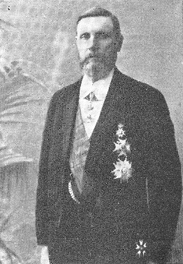 Ein Mann in einem schwarzen und weißen Anzug mit einem weißen Hemd und schwarzer Krawatte steht mit ernster Miene vor einer üppigen grünen Pflanze.