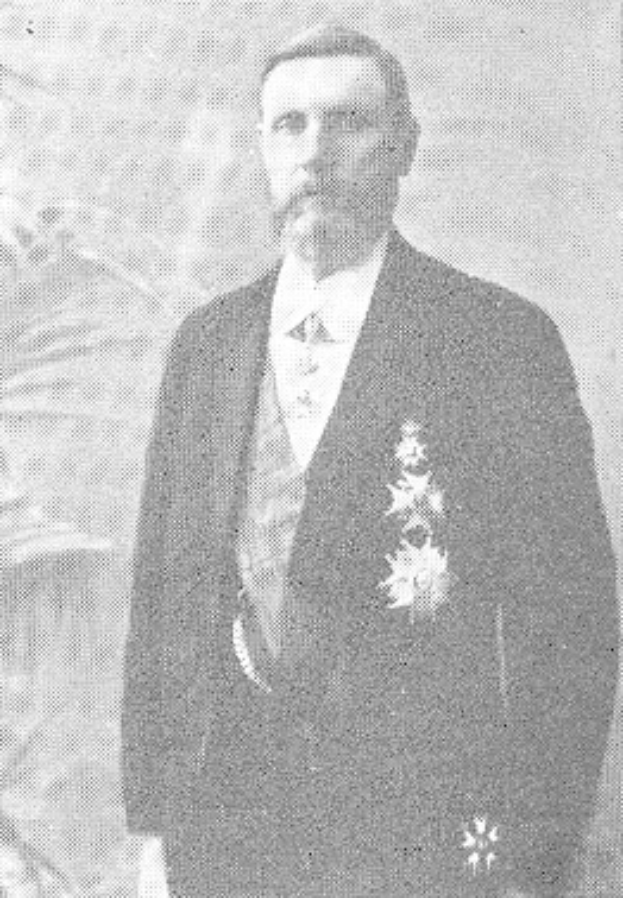 Ein Mann in einem schwarzen und weißen Anzug mit einem weißen Hemd und schwarzer Krawatte steht mit ernster Miene vor einer üppigen grünen Pflanze.