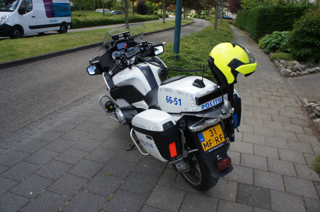 Ein auf der Seite geparktes Polizeimotorrad mit einem Helm darauf, mit Bäumen, Pfählen, Gebäuden, Pflanzen, Gras, Steinen und anderen Fahrzeugen im Hintergrund.