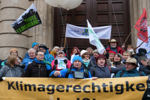 Eine Gruppe von Menschen steht vor einem Gebäude mit Säulen und einer Tür, hält eine Fahne mit Text, Fahnenstangen und Luftballons und nimmt an einem Klimastreik in Deutschland teil.