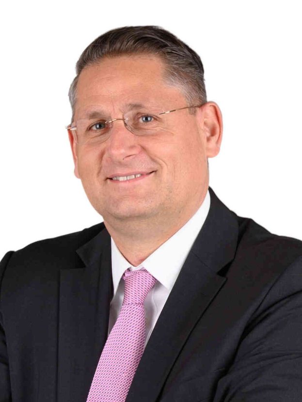 Ein lachender Mann im Anzug und Krawatte mit Brille vor einem weißen Hintergrund.