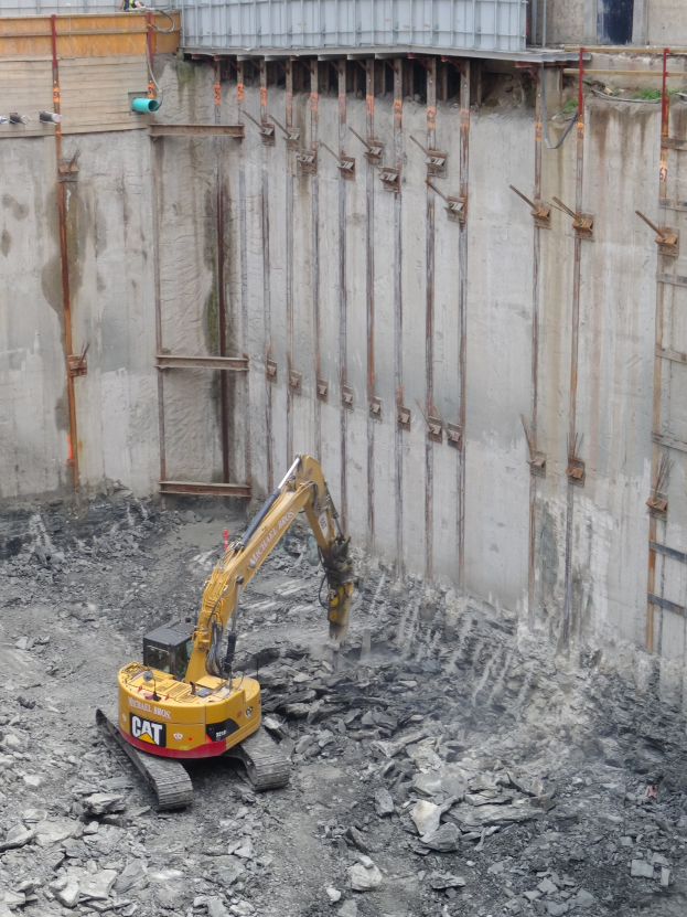 Bagger bei der Arbeit auf einer Baustelle mit Steinen und Eisenstangen neben einer teilweise errichteten Struktur.