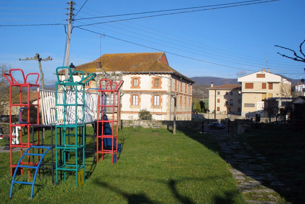 Ein Spielplatz in einer grünen Fläche, umgeben von Gebäuden, Bäumen, Strommasten, Fahrzeugen, Hügeln und einem klaren blauen Himmel.