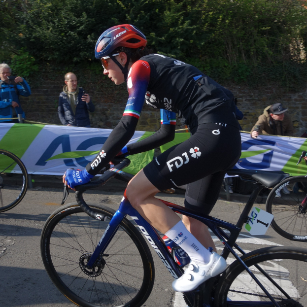 Eine professionelle Radfahrerin, Katie Archibald, trägt einen Helm, eine Schutzbrille, Handschuhe und Schuhe und fährt ein Fahrrad eine Straße entlang neben einer Menge Menschen, mit einem Banner, einer Wand, Bäumen und einem Gebäude im Hintergrund.