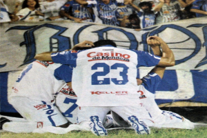 Eine Gruppe von Fußballspielern feiert ein Tor auf dem Platz vor einer jubelnden Menge, mit einem Banner mit Text im Hintergrund.