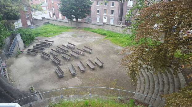 Ein leeres Amphitheater in einer Stadt, umgeben von Bäumen, Pflanzen, Gras, Treppen, Geländern, Pfosten und Gebäuden mit Fenstern, unter einem sichtbaren Himmel.