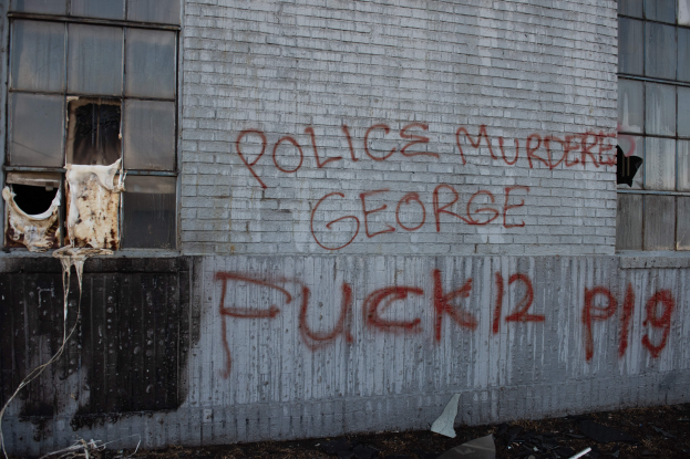 Ein Gebäude mit Graffiti, das "Polizei Mord George Fuck 12 P19" auf der Seite trägt, mit Fenstern und verschiedenen Gegenständen vor ihm.