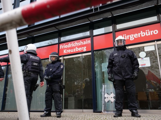 Eine Gruppe von Polizisten in Helmen und Handschuhen steht vor einem Glaswandgebäude, mit einem Pfahl links und Schildern im Hintergrund, die "Friedrichshain Kreuzberg" und Papiere an den Glaswänden angeklebt zeigen.