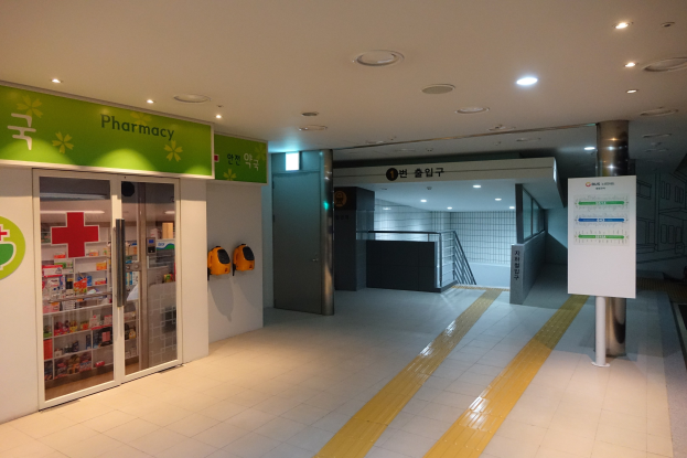 Eingang einer Apotheke mit einem "Apotheke"-Schild links, einer Tafel mit Text rechts, beleuchteten Lampen oben und Hintergrundobjekten.