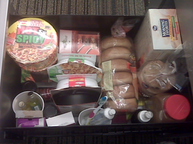 Ein Nahaufnahme einer Box mit verschiedenen Gegenständen, darunter Flaschen, eine Zahnbürste, kleinere Boxen und einige Lebensmittel.