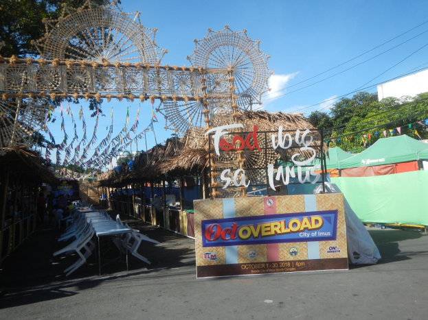 Eine KarnevalsSzene mit einem großen Riesenrad in der Mitte, umgeben von Hütten, Sitzbereichen, einer Tafel, Dekorationen, Bäumen, Überlandleitungen und einem bewölkten Himmel.