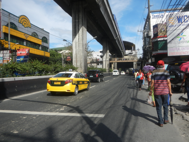 Ein gelbes Taxi fährt auf einer Stadtstraße mit hohen Gebäuden, Fußgängern mit Schirmen, einer Brücke im Hintergrund und bewölktem Himmel.