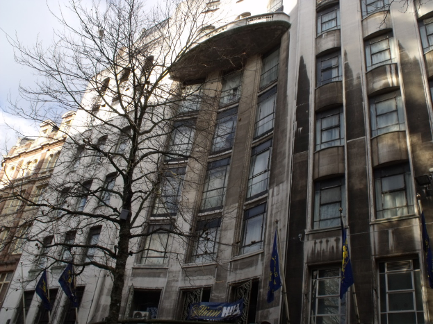 Gebäude mit Fenstern, einer Fahne und Flaggen an Stangen unten sowie einem Baum mit Zweigen.