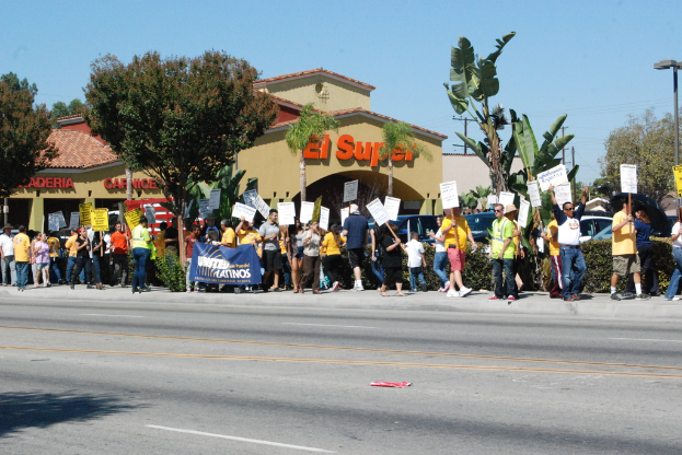 Eine Gruppe von Menschen mit Schildern marschiert auf einer Straße mit Bäumen, Büschen und Gebäuden mit Namensschildern, Autos und einem Banner im Hintergrund.