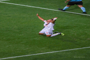 Ein Fußballspieler kniet auf dem Boden mit ausgestreckten Armen in Feierlaune, trägt eine Sportuniform und Schuhe.