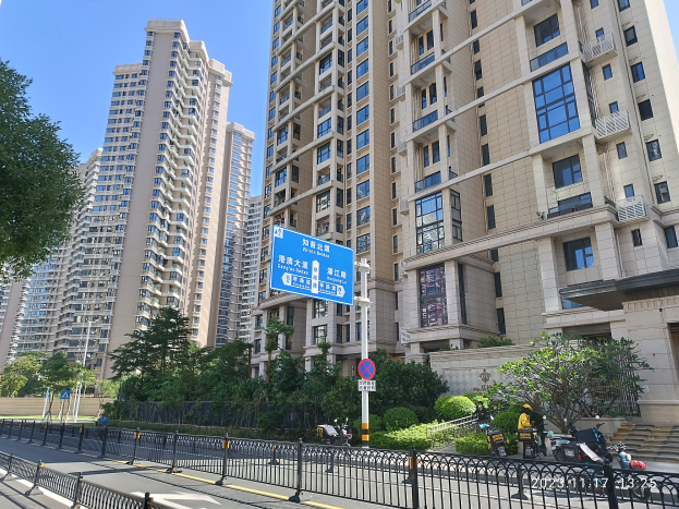 Eine Straßenansicht in Hong Kong mit hohen Wohngebäuden, Bäumen, Pflanzen, einem Zaun, Schildern, Pfosten, Fahrzeugen und einem klaren blauen Himmel.