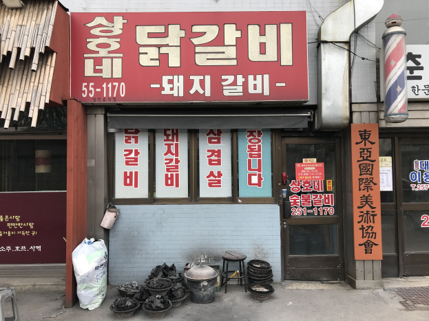 Außenansicht eines Barbiershops in Seoul, Südkorea, mit einem Schild, Hockern, Eimern, einem Müllbehälter, einer Abdeckung, einem Pfahl mit einer Platte und Glasfenstern und einer Tür mit Texttafeln.
