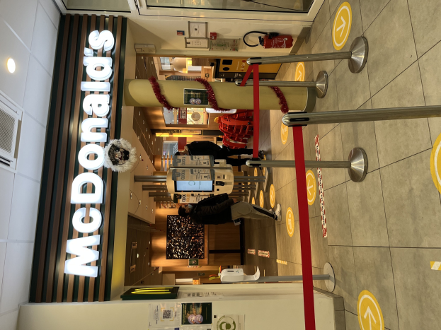 Innere eines McDonald's-Restaurants mit einem sichtbaren 'McDonald's'-Schild auf der linken Seite, roten Metallstangen mit Bändern auf der rechten Seite und einigen Menschen in der Nähe des Eingangs mit Hintergrundobjekten.